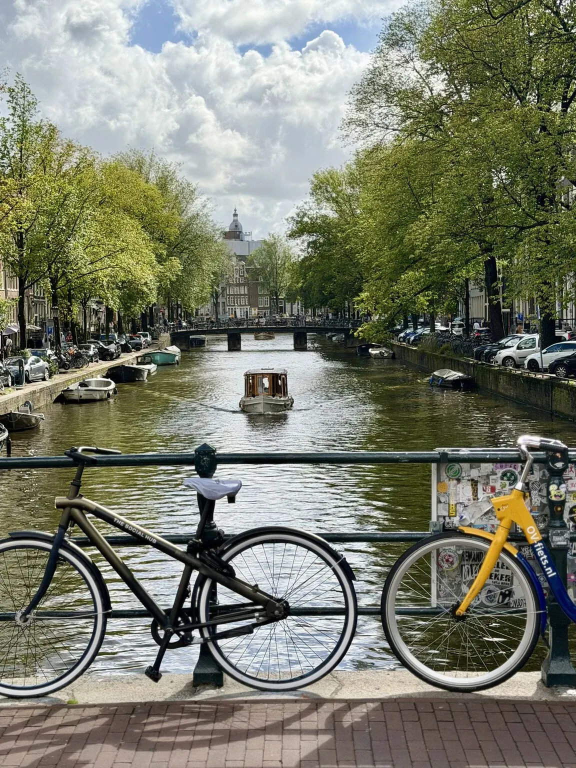 Tree-lined Amsterdam canal with a small boat, seen from a bridge with two bicycles against the railing